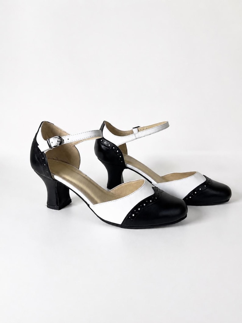 May include: A pair of black and white two-tone shoes with a rounded toe, a mid-height block heel, and an ankle strap featuring a silver-toned metal buckle. The design includes decorative perforations along the wingtip-style toe cap seam.