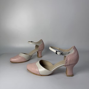 Op de afbeelding: Een paar tweekleurige schoenen in een lichtroze en cr&egrave;mekleurig schema. Deze schoenen hebben een ronde neus, een halflange uitlopende hak en een verstelbare enkelband met een zilverkleurige metalen gesp. Het ontwerp bevat decoratieve perforaties in brogue-stijl langs de naden.