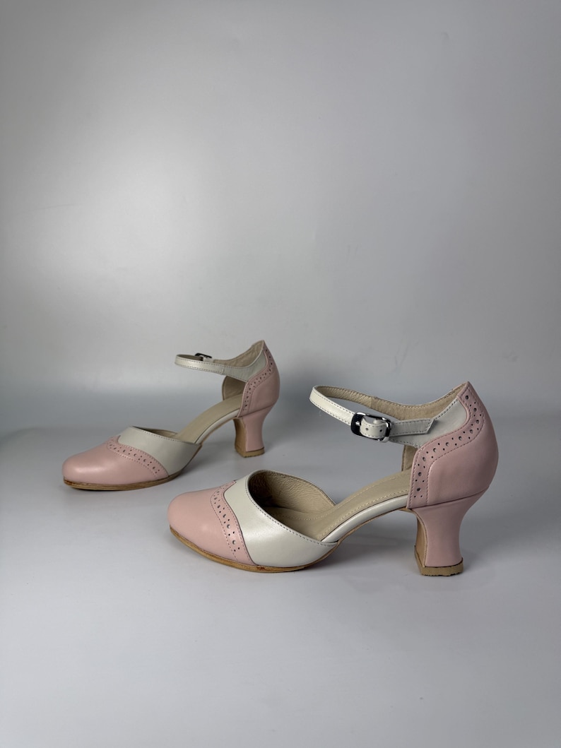 Op de afbeelding: Een paar tweekleurige schoenen in een lichtroze en cr&egrave;mekleurig schema. Deze schoenen hebben een ronde neus, een halflange uitlopende hak en een verstelbare enkelband met een zilverkleurige metalen gesp. Het ontwerp bevat decoratieve perforaties in brogue-stijl langs de naden.