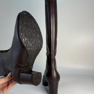 May include: A pair of tall, dark brown leather boots. One boot is angled to show the sole with a detailed pattern, while the other stands upright. Both boots have a block heel and a smooth, polished finish, suggesting a classic design.