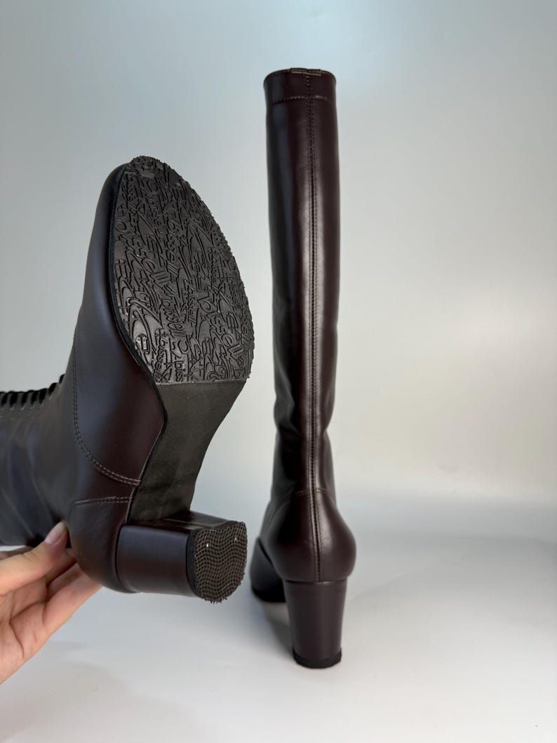 May include: A pair of tall, dark brown leather boots. One boot is angled to show the sole with a detailed pattern, while the other stands upright. Both boots have a block heel and a smooth, polished finish, suggesting a classic design.