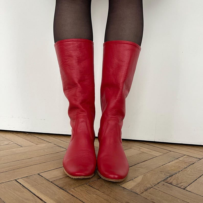 Pode incluir: Um par de botas altas de couro vermelho. As botas t&ecirc;m uma textura suave e um design cl&aacute;ssico, com bico arredondado e altura a meio da panturrilha. As botas est&atilde;o sobre um ch&atilde;o de madeira.