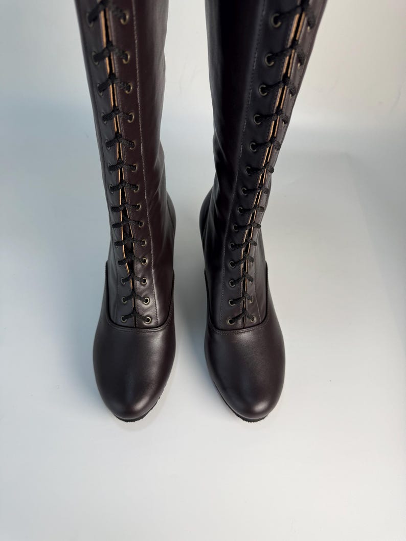 May include: A pair of dark brown knee-high boots with a lace-up design. The boots have a rounded toe and a subtle heel. The laces are black, and the boots appear to be made of leather.