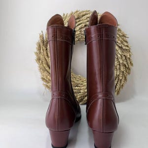 May include: A pair of mid-calf brown leather boots featuring a smooth finish, a low block heel, and decorative stitching details with small circular perforations across the upper shaft and near the heel. The boots have a side zipper closure.