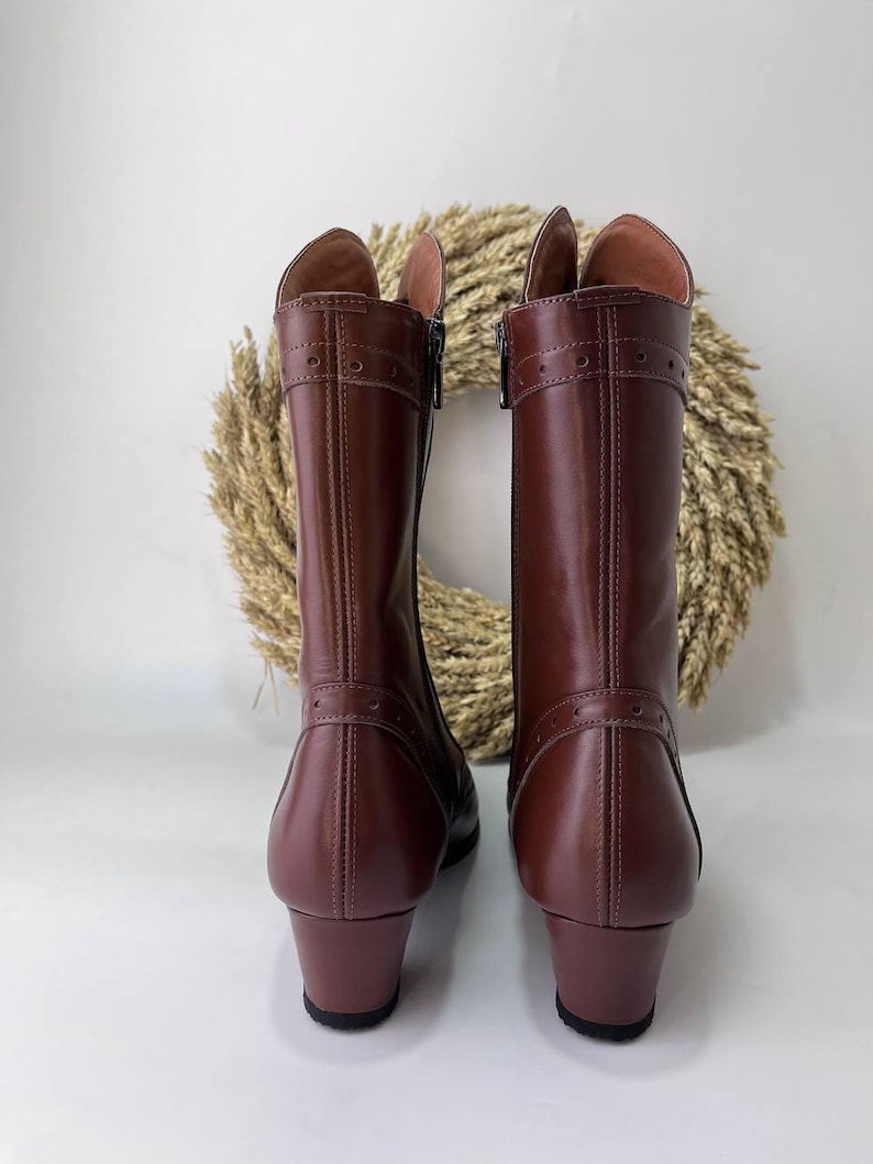 May include: A pair of mid-calf brown leather boots featuring a smooth finish, a low block heel, and decorative stitching details with small circular perforations across the upper shaft and near the heel. The boots have a side zipper closure.