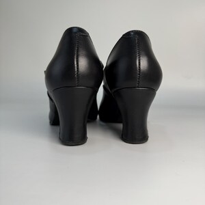 May include: A pair of black leather pumps with a rounded toe and a mid-height, curved heel. The shoes have a classic design, suitable for formal or professional settings. The heels are approximately 2.5 inches (6.35 cm) high.