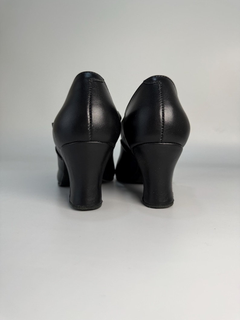 May include: A pair of black leather pumps with a rounded toe and a mid-height, curved heel. The shoes have a classic design, suitable for formal or professional settings. The heels are approximately 2.5 inches (6.35 cm) high.