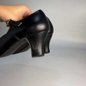 May include: Black leather shoes with a small heel. The shoes have a strap with a buckle closure. The heel is approximately 2 inches high. The shoes are held up by a hand.