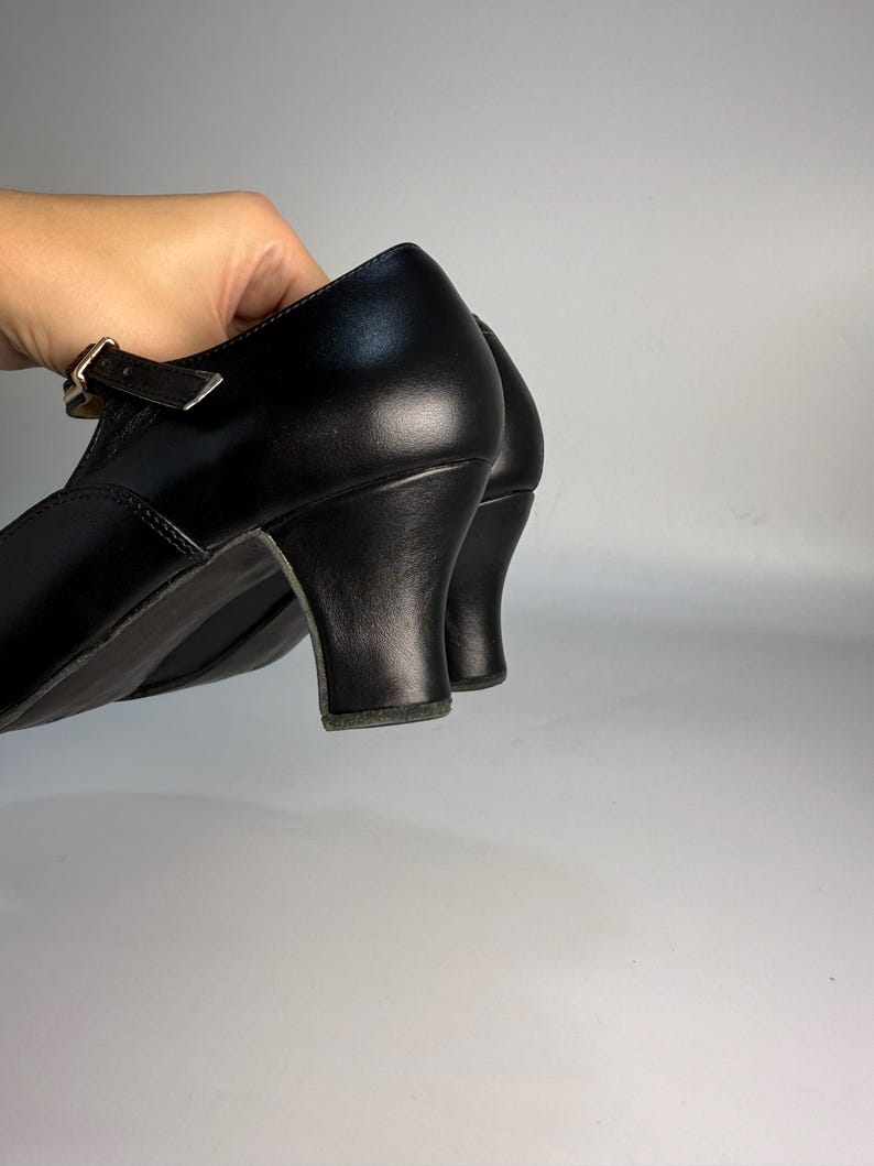 May include: Black leather shoes with a small heel. The shoes have a strap with a buckle closure. The heel is approximately 2 inches high. The shoes are held up by a hand.