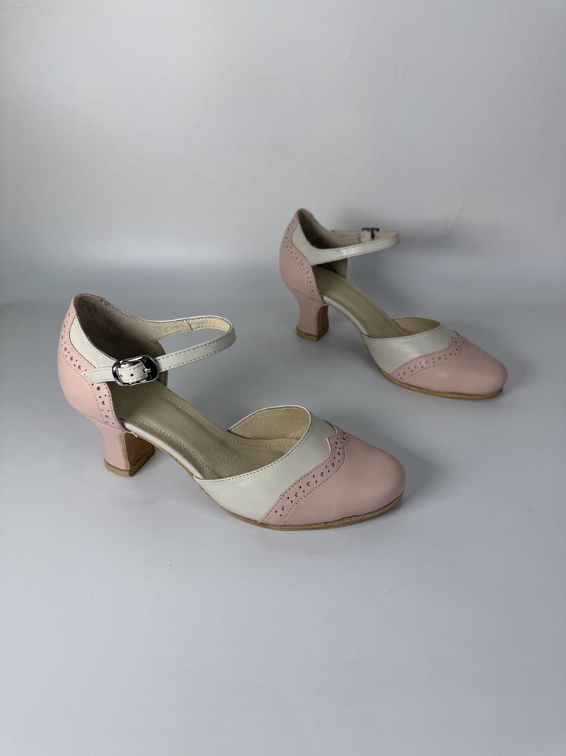 Op de afbeelding: Een paar tweekleurige Mary Jane-hakken in lichtroze en cr&egrave;me. Deze schoenen hebben een ronde neus, een lage blokhak en een verstelbare enkelband met een zilverkleurige metalen gesp. Het ontwerp bevat decoratieve perforaties in brogue-stijl.
