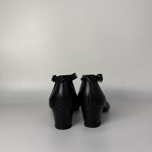 May include: A pair of black shoes with a low block heel and an ankle strap featuring a silver-toned metal buckle. The shoes have a smooth, solid black finish and are viewed from the back, showing the heel construction and the strap closure.