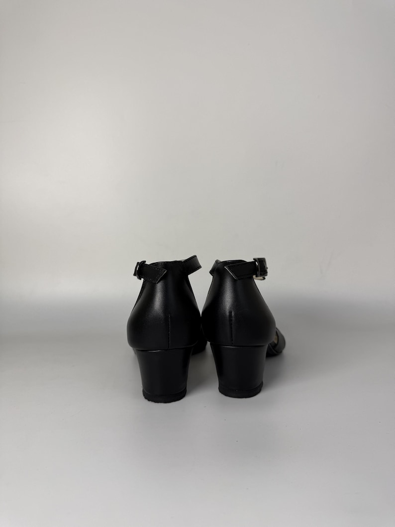 May include: A pair of black shoes with a low block heel and an ankle strap featuring a silver-toned metal buckle. The shoes have a smooth, solid black finish and are viewed from the back, showing the heel construction and the strap closure.