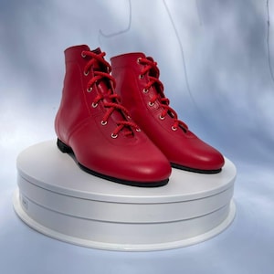 May include: A pair of vibrant red ankle boots with red laces. The boots feature a classic design with a rounded toe and a low heel. They are presented on a white, rotating platform against a light blue backdrop.