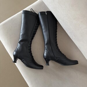 May include: A pair of black knee-high boots featuring a lace-up front design, a side zipper closure, and a low, curved heel. The boots are detailed with decorative perforations along the seams and a smooth finish throughout the shaft and toe.