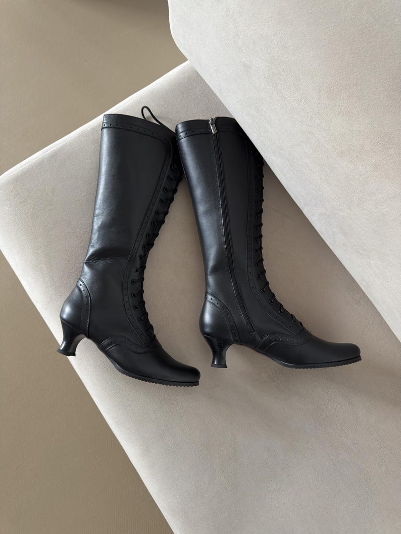 May include: A pair of black knee-high boots featuring a lace-up front design, a side zipper closure, and a low, curved heel. The boots are detailed with decorative perforations along the seams and a smooth finish throughout the shaft and toe.