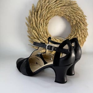 Puede incluir: Un par de zapatos de tac&oacute;n negros con tira en T, acabado en cuero liso, punta redondeada y correa al tobillo con hebilla. Los zapatos tienen un tac&oacute;n curvo de altura media y un forro interior claro, con detalles de aberturas en los laterales.