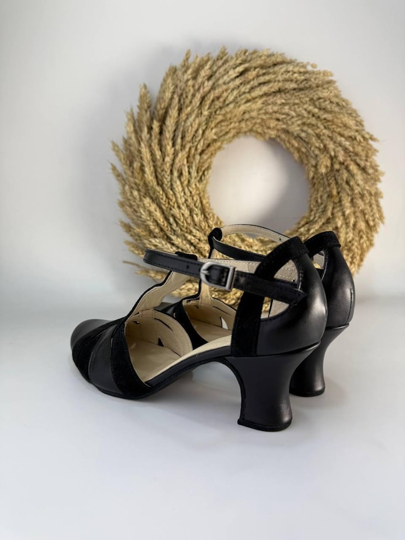 Puede incluir: Un par de zapatos de tac&oacute;n negros con tira en T, acabado en cuero liso, punta redondeada y correa al tobillo con hebilla. Los zapatos tienen un tac&oacute;n curvo de altura media y un forro interior claro, con detalles de aberturas en los laterales.