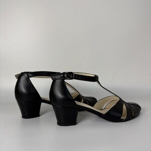May include: A pair of black T-strap shoes with a low block heel and an adjustable ankle strap featuring a metal buckle. The shoes have a closed toe with cut-out details on the sides and a light-coloured interior lining visible at the heel and toe.