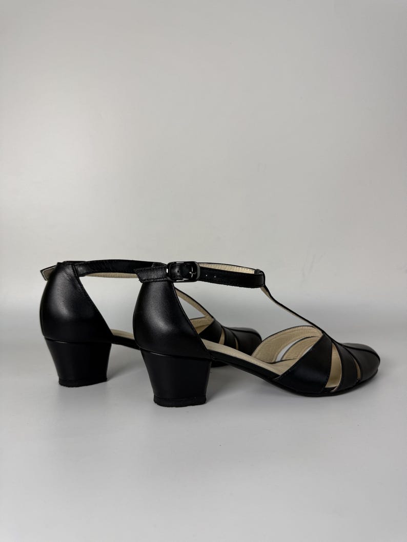 May include: A pair of black T-strap shoes with a low block heel and an adjustable ankle strap featuring a metal buckle. The shoes have a closed toe with cut-out details on the sides and a light-coloured interior lining visible at the heel and toe.