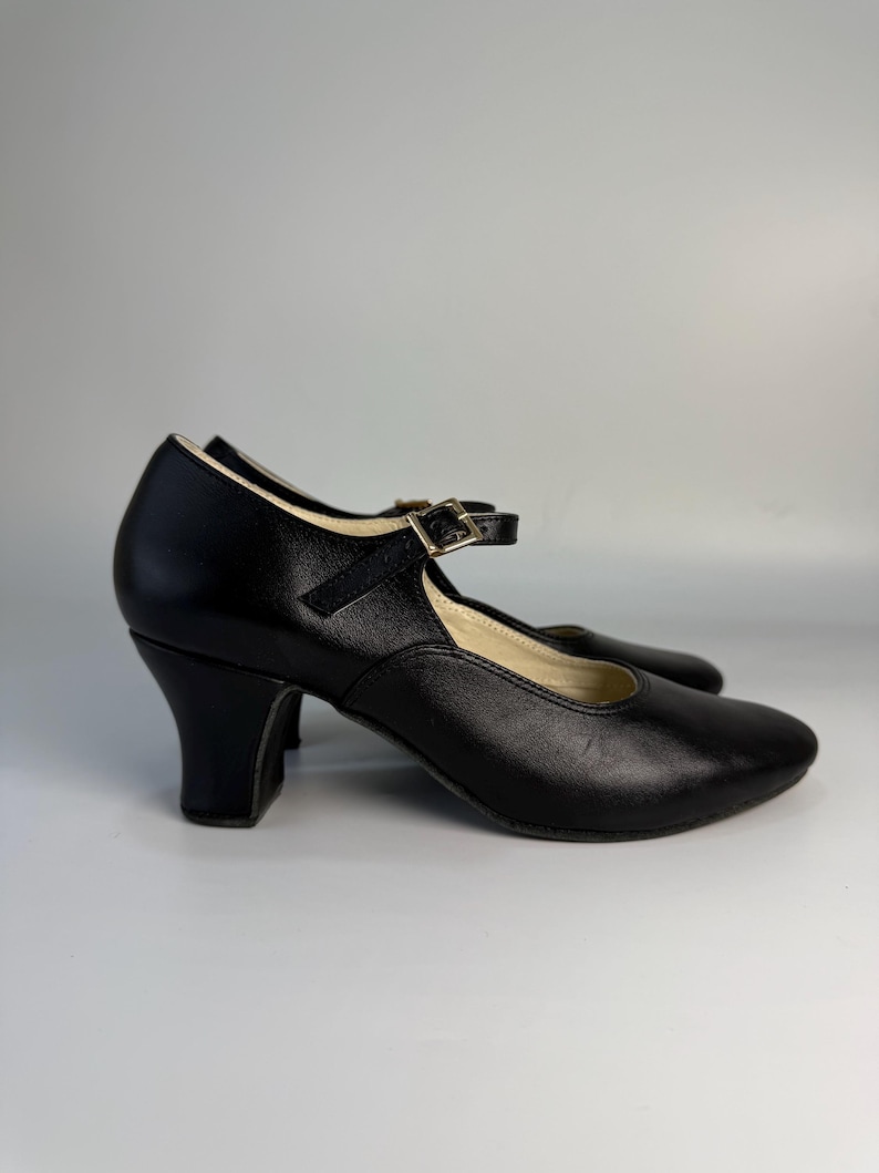 May include: Black leather Mary Jane style shoes with a strap and buckle closure. The shoes have a rounded toe and a small, curved heel. The shoes are set against a plain white background.