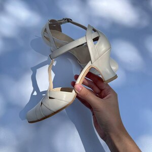 Puede incluir: Un par de zapatos de baile con tira en T de color blanco roto, con puntera redondeada, tac&oacute;n acampanado de altura media y correa al tobillo con hebilla. Los zapatos presentan tiras entrecruzadas en el empeine y los laterales para mayor sujeci&oacute;n.