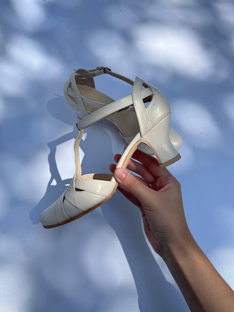 Puede incluir: Un par de zapatos de baile con tira en T de color blanco roto, con puntera redondeada, tac&oacute;n acampanado de altura media y correa al tobillo con hebilla. Los zapatos presentan tiras entrecruzadas en el empeine y los laterales para mayor sujeci&oacute;n.