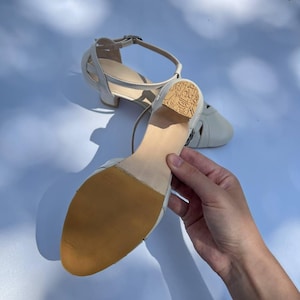 Puede incluir: Un par de zapatos de baile de cuero color blanco roto con correa al tobillo y tac&oacute;n bajo de bloque. La suela es de color tostado y texturizada, mientras que el tac&oacute;n presenta un patr&oacute;n grabado. Tienen punta redondeada y cierre de hebilla.