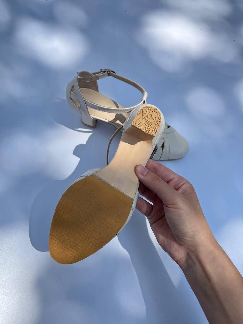 Puede incluir: Un par de zapatos de baile de cuero color blanco roto con correa al tobillo y tac&oacute;n bajo de bloque. La suela es de color tostado y texturizada, mientras que el tac&oacute;n presenta un patr&oacute;n grabado. Tienen punta redondeada y cierre de hebilla.
