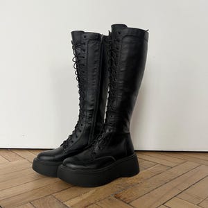May include: A pair of black knee-high platform boots. The boots have a lace-up design and a thick sole. The boots are made of a smooth, leather-like material. The boots are displayed on a wooden floor against a white wall.