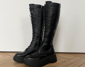 May include: A pair of black knee-high platform boots. The boots feature a lace-up design and a thick sole. The boots are made of a smooth, leather-like material. The boots are displayed on a wooden floor against a white wall.