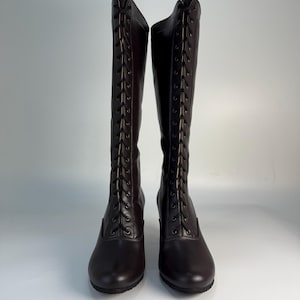 May include: A pair of dark brown, knee-high boots. The boots have a lace-up design on the front and a small heel. The boots are made of a smooth material and are displayed against a white background.