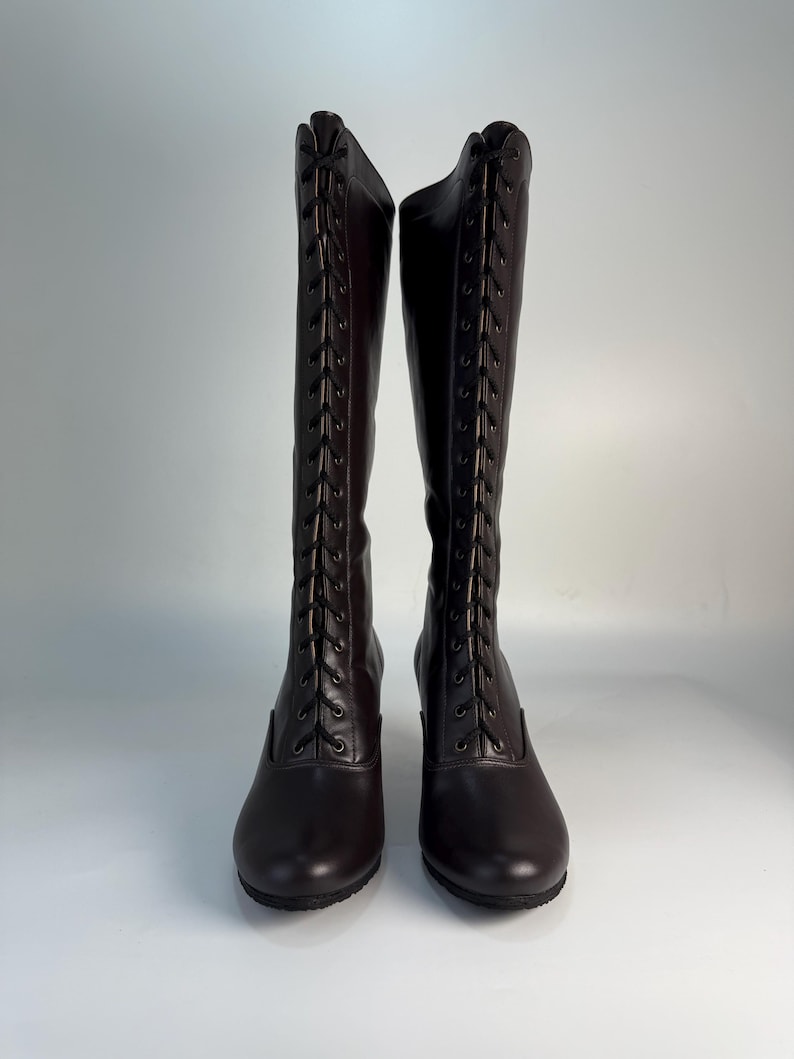 May include: A pair of dark brown, knee-high boots. The boots have a lace-up design on the front and a small heel. The boots are made of a smooth material and are displayed against a white background.