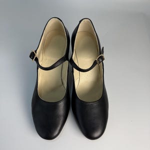 May include: A pair of black Mary Jane style shoes with a rounded toe and a strap that fastens with a gold-colored buckle. The shoes have a low heel and a smooth leather-like appearance. The interior of the shoe is a light beige color.