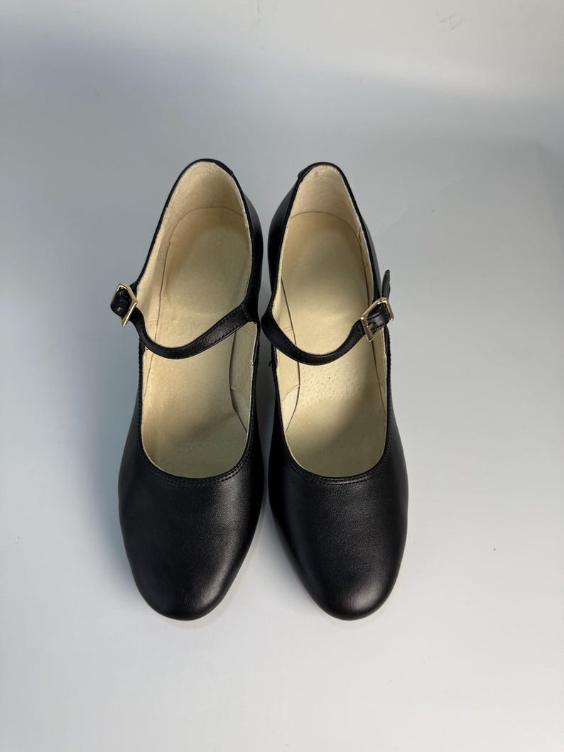 May include: A pair of black Mary Jane style shoes with a rounded toe and a strap that fastens with a gold-colored buckle. The shoes have a low heel and a smooth leather-like appearance. The interior of the shoe is a light beige color.