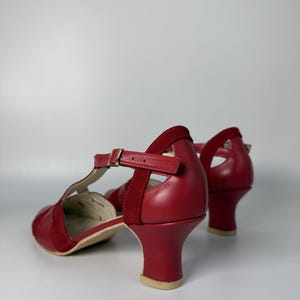 Puede incluir: Un par de zapatos de pulsera en T rojos con acabado en piel lisa y detalles en ante. Los zapatos tienen puntera redondeada, tac&oacute;n de bloque de altura media y una correa ajustable en el tobillo con una peque&ntilde;a hebilla de metal dorado.