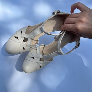 Puede incluir: Un par de zapatos de cuero color blanco hueso con punta redondeada, dise&ntilde;o recortado en el empeine y correa de tobillo ajustable con hebilla de metal plateado. Los zapatos tienen un tac&oacute;n bajo de bloque y un acabado liso de color s&oacute;lido.