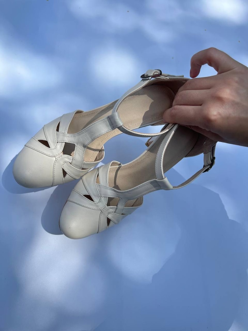 Puede incluir: Un par de zapatos de cuero color blanco hueso con punta redondeada, dise&ntilde;o recortado en el empeine y correa de tobillo ajustable con hebilla de metal plateado. Los zapatos tienen un tac&oacute;n bajo de bloque y un acabado liso de color s&oacute;lido.