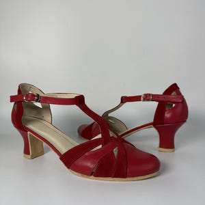 Puede incluir: Un par de zapatos rojos con tira en T que combinan texturas de cuero liso y ante. Los zapatos tienen la punta redondeada, un tac&oacute;n de bloque de altura media con base clara y una correa ajustable en el tobillo con una peque&ntilde;a hebilla dorada.
