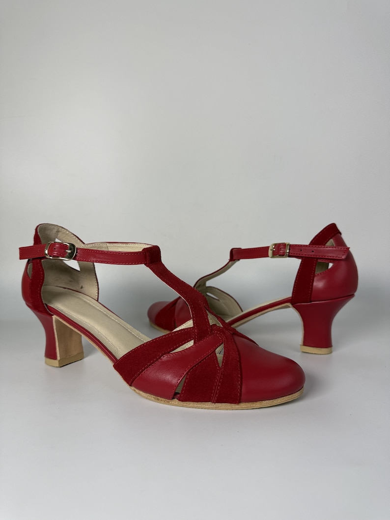 Puede incluir: Un par de zapatos rojos con tira en T que combinan texturas de cuero liso y ante. Los zapatos tienen la punta redondeada, un tac&oacute;n de bloque de altura media con base clara y una correa ajustable en el tobillo con una peque&ntilde;a hebilla dorada.