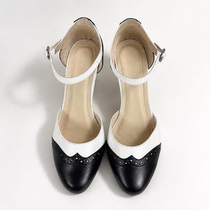 May include: A pair of two-tone Mary Jane shoes featuring a black rounded toe with decorative brogue perforations, a white mid-section, and a white ankle strap with a small silver-toned buckle. The interior lining is a light beige color.