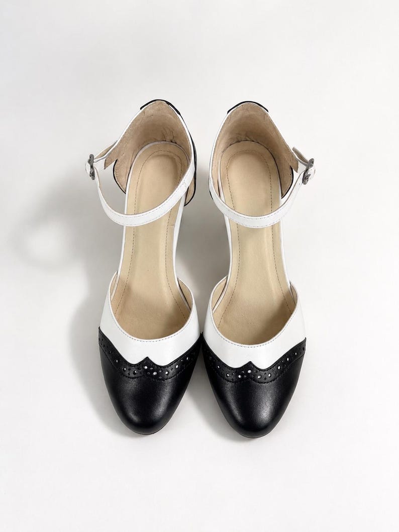May include: A pair of two-tone Mary Jane shoes featuring a black rounded toe with decorative brogue perforations, a white mid-section, and a white ankle strap with a small silver-toned buckle. The interior lining is a light beige color.
