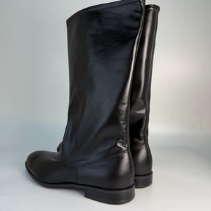 Puede incluir: Un par de botas negras de media caña con acabado liso y mate, punta redondeada y tacón bajo y plano. Las botas tienen una caña estructurada que llega hasta la mitad de la pantorrilla, con costuras verticales visibles en la parte posterior.