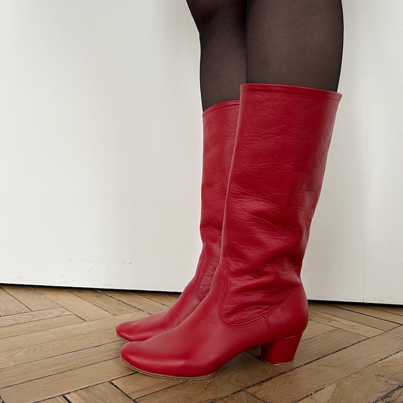 Pode incluir: Um par de botas altas de couro vermelho, com bico arredondado e salto pequeno. As botas t&ecirc;m uma textura lisa e um design cl&aacute;ssico. As botas chegam aproximadamente ao joelho.