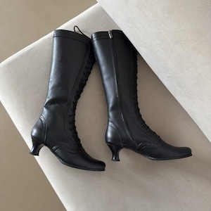 A pair of black knee-high boots featuring a lace-up front closure, a side zipper, and a small kitten heel. The boots are detailed with decorative brogue-style perforations along the toe cap, heel counter, and the vertical lace panel.