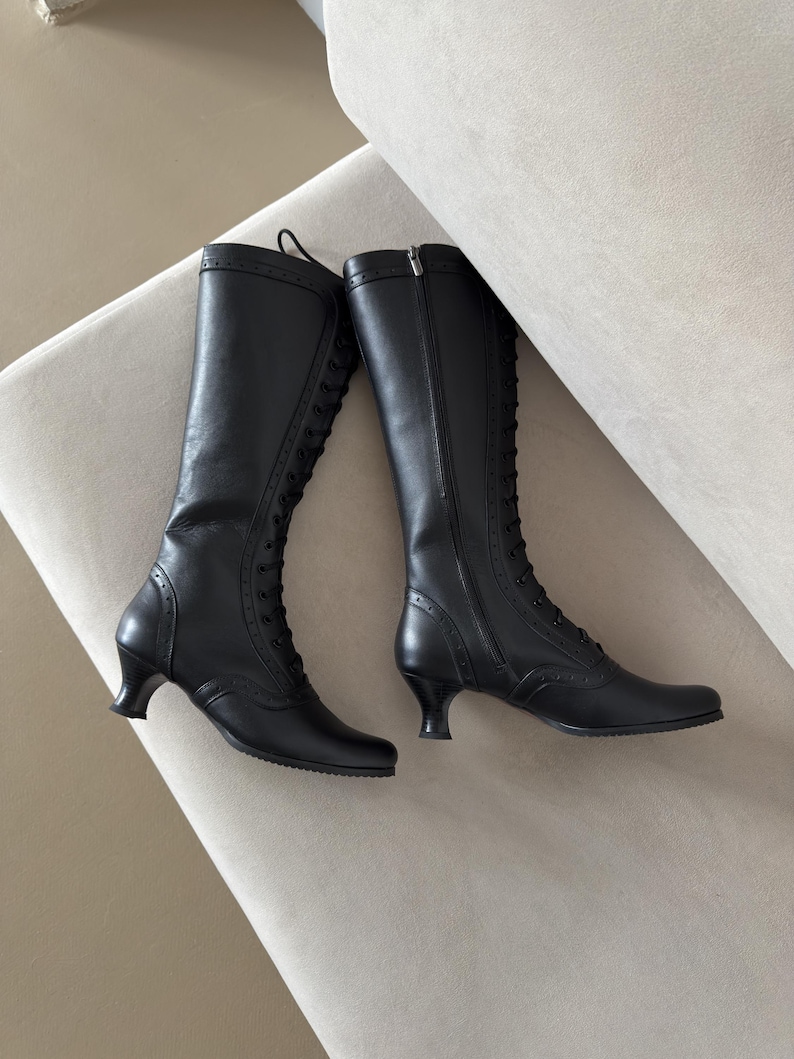 A pair of black knee-high boots featuring a lace-up front closure, a side zipper, and a small kitten heel. The boots are detailed with decorative brogue-style perforations along the toe cap, heel counter, and the vertical lace panel.
