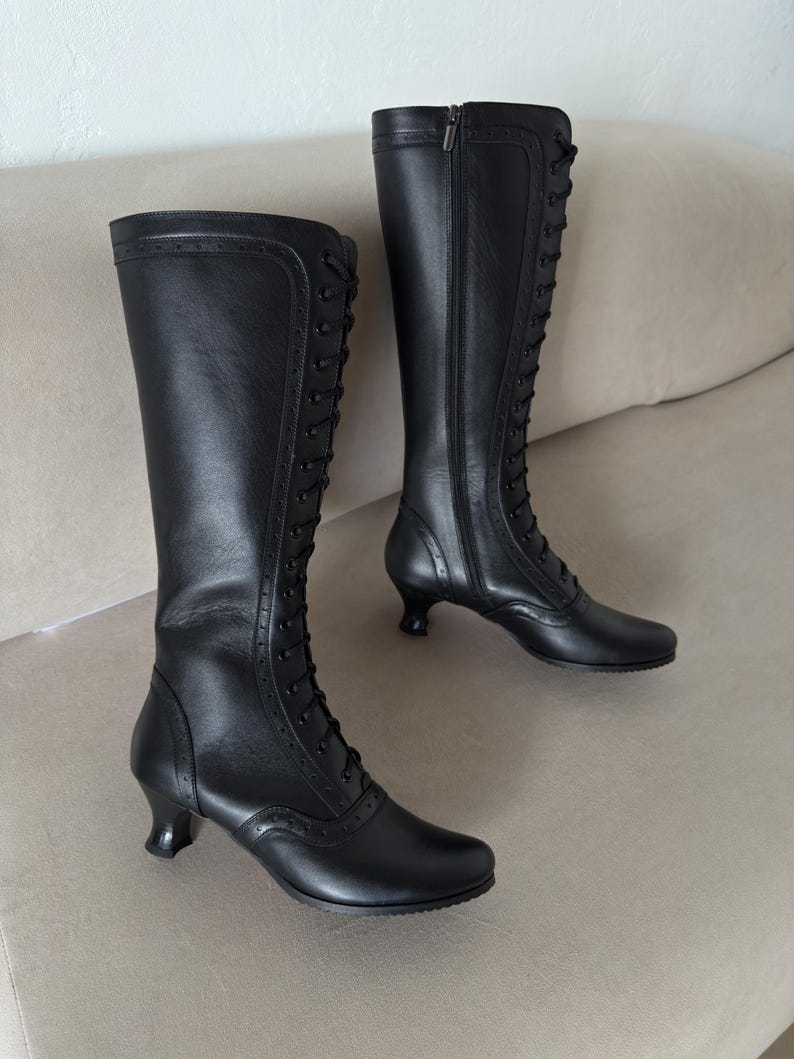 May include: A pair of black leather knee-high boots featuring a lace-up front with black laces, decorative brogue-style perforated detailing along the seams, a side zipper closure, and a small, flared kitten heel for a structured appearance.