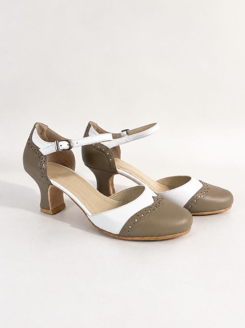 May include: A pair of two-tone taupe and white heels featuring a rounded toe, a cutout side design, and a thin ankle strap with a silver-toned metal buckle. The shoes include decorative brogue-style perforations along the toe and heel sections.