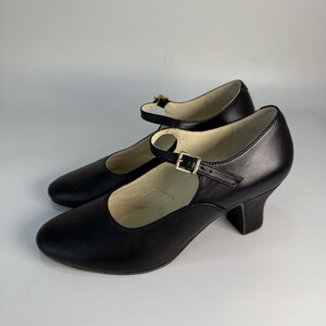 May include: A pair of black leather Mary Jane style shoes with a rounded toe and a small heel. Each shoe has a strap that fastens with a gold-colored buckle. The shoes are set against a white background.