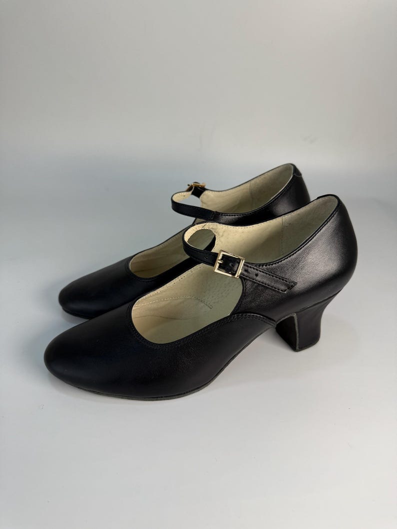 May include: A pair of black leather Mary Jane style shoes with a rounded toe and a small heel. Each shoe has a strap that fastens with a gold-colored buckle. The shoes are set against a white background.
