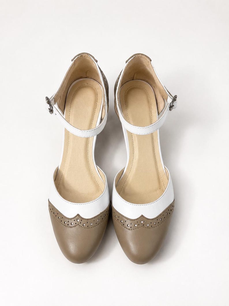 May include: A pair of two-tone Mary Jane shoes featuring a white upper section and a taupe-colored rounded toe cap. The shoes include a decorative brogue-style perforated trim along the seam, a thin ankle strap with a metal buckle, and a beige insole.